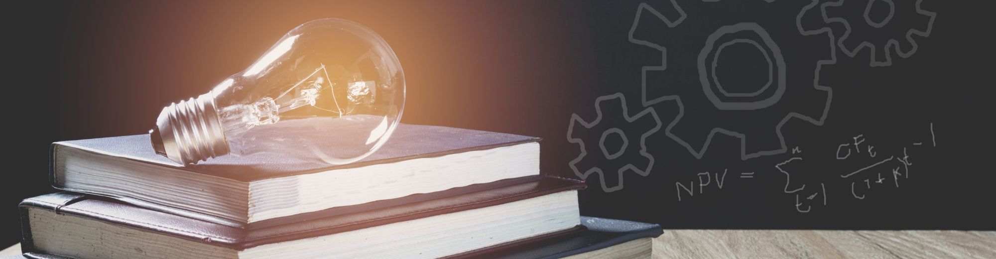 Glühbirne liegt auf zwei übereinander gestapelten Büchern vor einer Tafel mit skizzierten Zahnrädern und Formeln